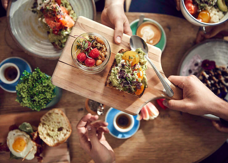 someone passing brekkie board to friend across the table, with avo smash and coffees on table. 