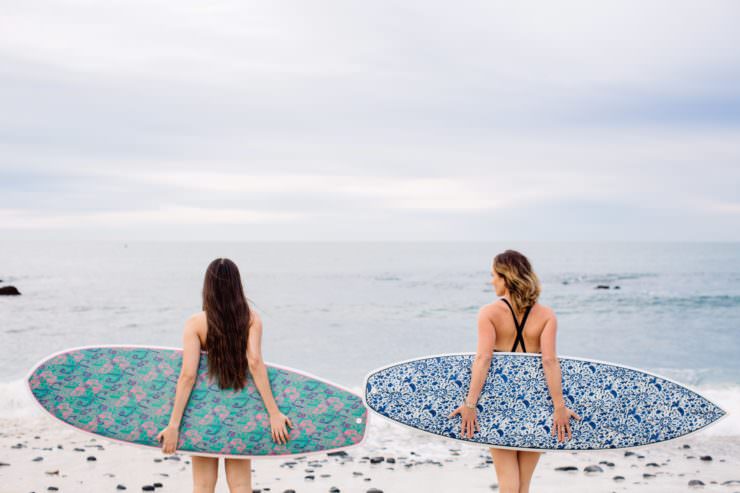 Two girls holding surfboards behind their backs on the beach. 