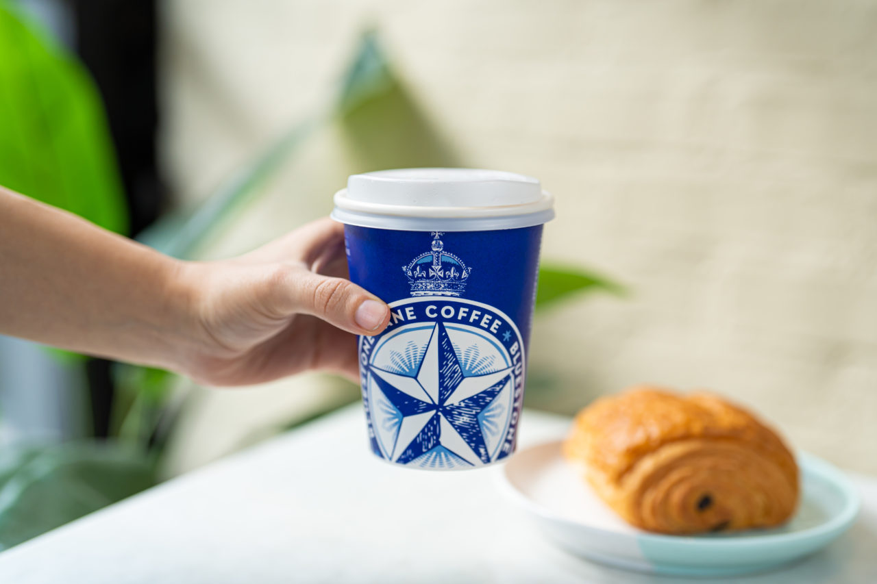 A hand holding a Bluestone Lane takeaway cup with a croissant in the background