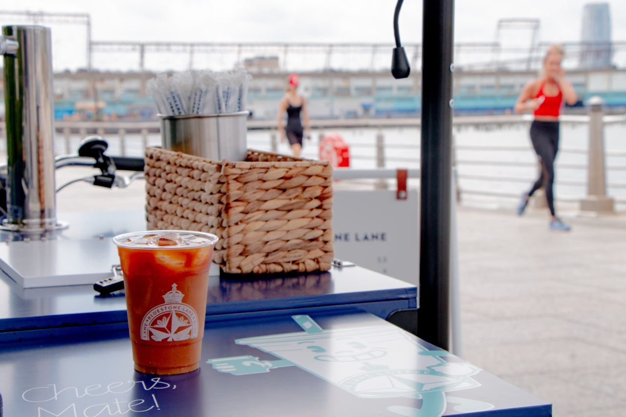 A Bluestone Lane takeaway cup with an iced drink inside, sitting on a takeaway counter with a running path in the background