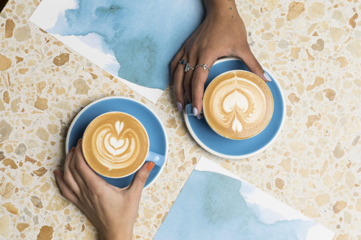 A birds eye view of two hands, each holding a flat white coffee