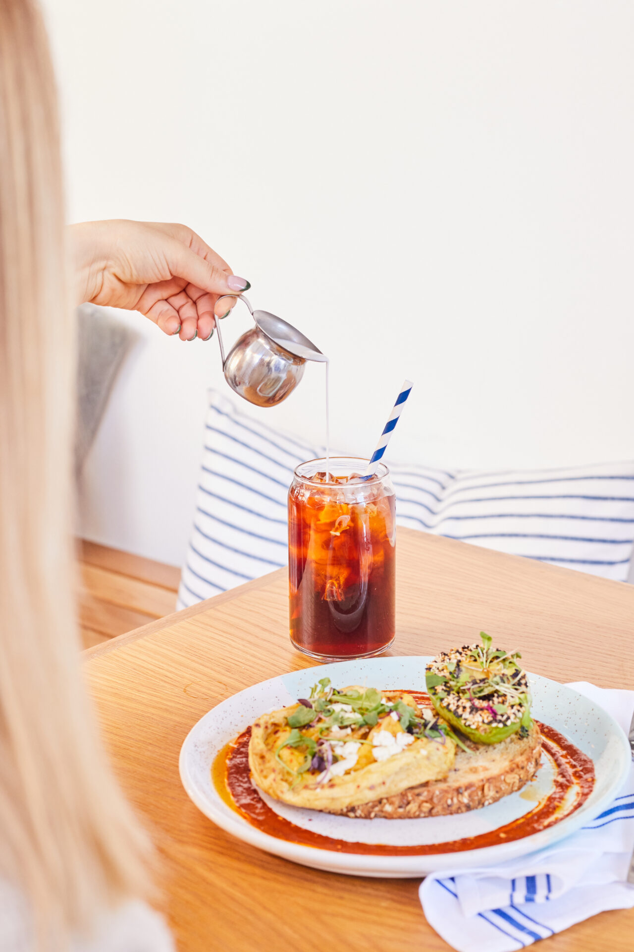 A women pours milk into a cold brew with a plate of scrambled egg and toast on the table.