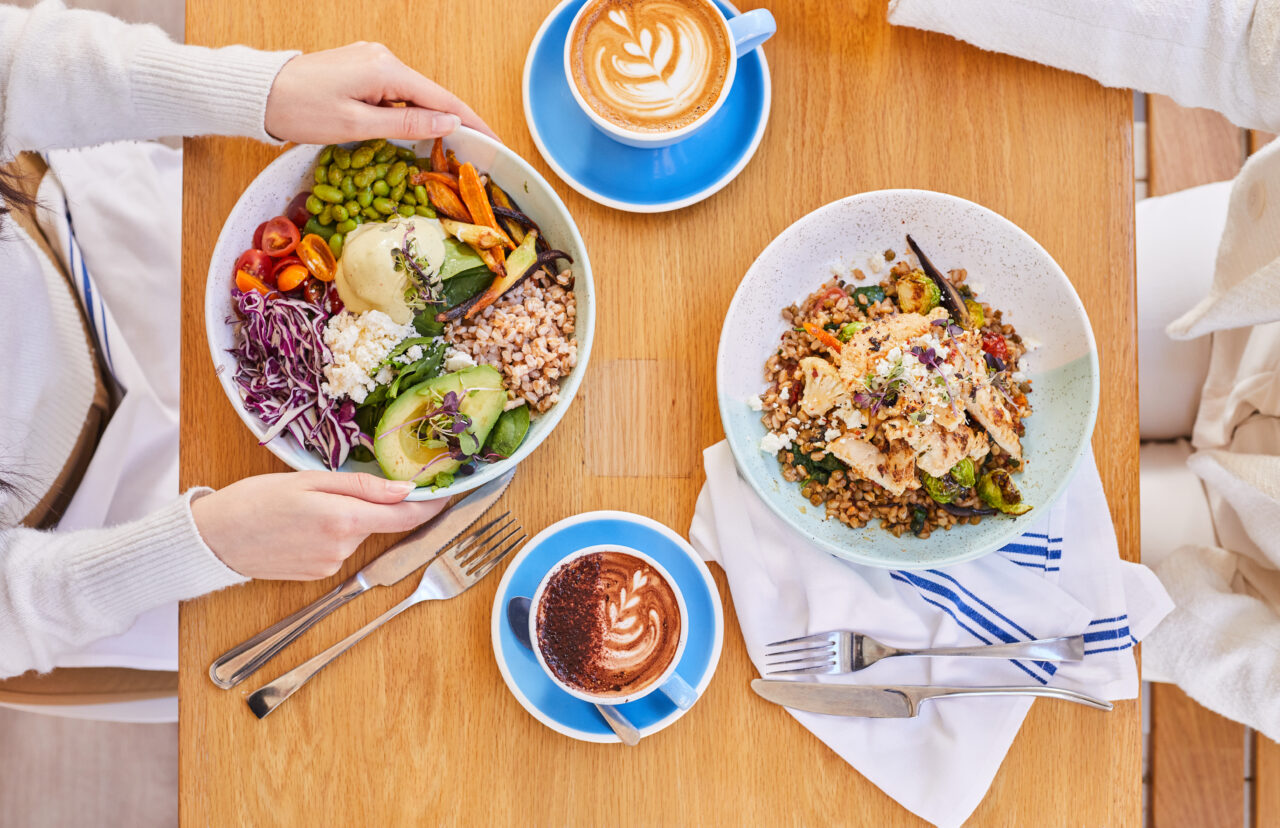 A birds eye view of a brunch table with Bluestone Lane salads, a hot chocolate and flat white.