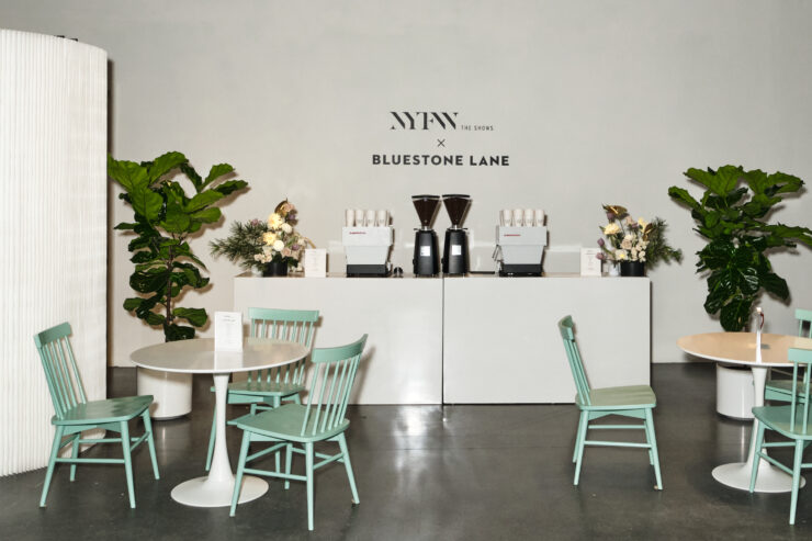 Bluestone Lane x NYFW set up - teal chairs are positiobed around white tables. There is a coffee station in the background and a wall decal with the words NYFW x Bluestone Lane above it.